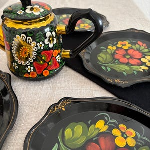 May include: A black, hand-painted teapot and serving trays with floral designs. The teapot has a black handle and lid, with gold trim. The trays are black with colorful flowers in red, yellow, and green. The set is on a neutral-colored surface.