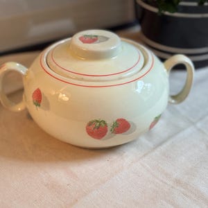 Peut inclure: Un sucrier en c&eacute;ramique de couleur cr&egrave;me avec deux anses lat&eacute;rales et un couvercle assorti. Le bol et le couvercle pr&eacute;sentent un motif r&eacute;p&eacute;titif de fraises rouges avec des feuilles vertes et de fines lignes rouges circulaires sur le bord.