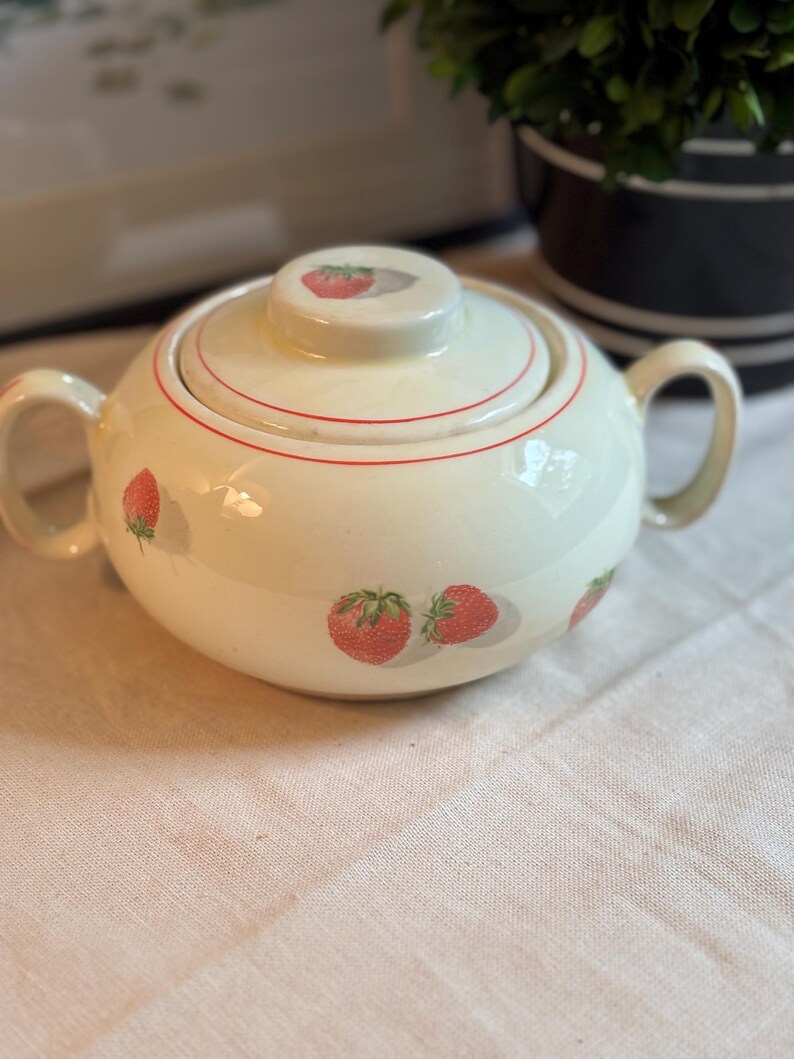 Peut inclure: Un sucrier en c&eacute;ramique de couleur cr&egrave;me avec deux anses lat&eacute;rales et un couvercle assorti. Le bol et le couvercle pr&eacute;sentent un motif r&eacute;p&eacute;titif de fraises rouges avec des feuilles vertes et de fines lignes rouges circulaires sur le bord.