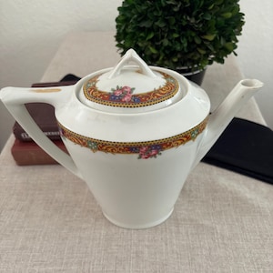 May include: White porcelain teapot with a lid, featuring a decorative band of red, gold, and green floral patterns. The teapot has a long spout and a curved handle. A small green plant is in the background.
