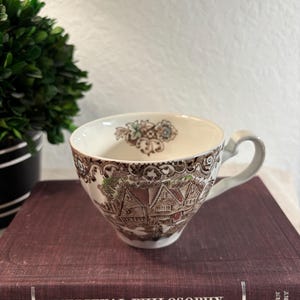 May include: A vintage teacup with a detailed brown and white transferware design depicting a village scene. The cup has a curved handle and is set on a stack of books. The books have the title "Medieval Philosophy".
