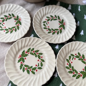 May include: Four off-white ceramic plates with a scalloped, fluted edge design. Each plate features a central circular illustration of a green holly leaf wreath with bright red berries. The plates are arranged on a dark green fabric surface.
