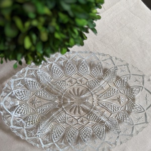 May include: An oval-shaped clear glass serving platter featuring a detailed pressed pattern of geometric shapes, starbursts, and cross-hatched textures. The dish has a scalloped edge and a central circular motif with radiating petal-like segments.