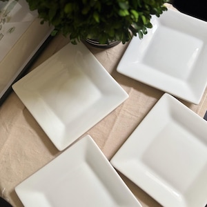 May include: A set of four white square ceramic plates with smooth, glossy surfaces and slightly raised edges. The plates are arranged in a cluster, showcasing their clean, minimalist design and uniform white finish for serving food.