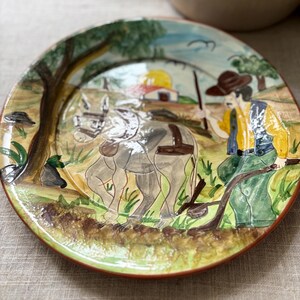 May include: A round ceramic plate featuring a hand-painted scene of a person in a brown hat and yellow shirt guiding a grey donkey through a field. The background includes a small building, a tree, and a yellow sun, all rendered in a glossy finish.
