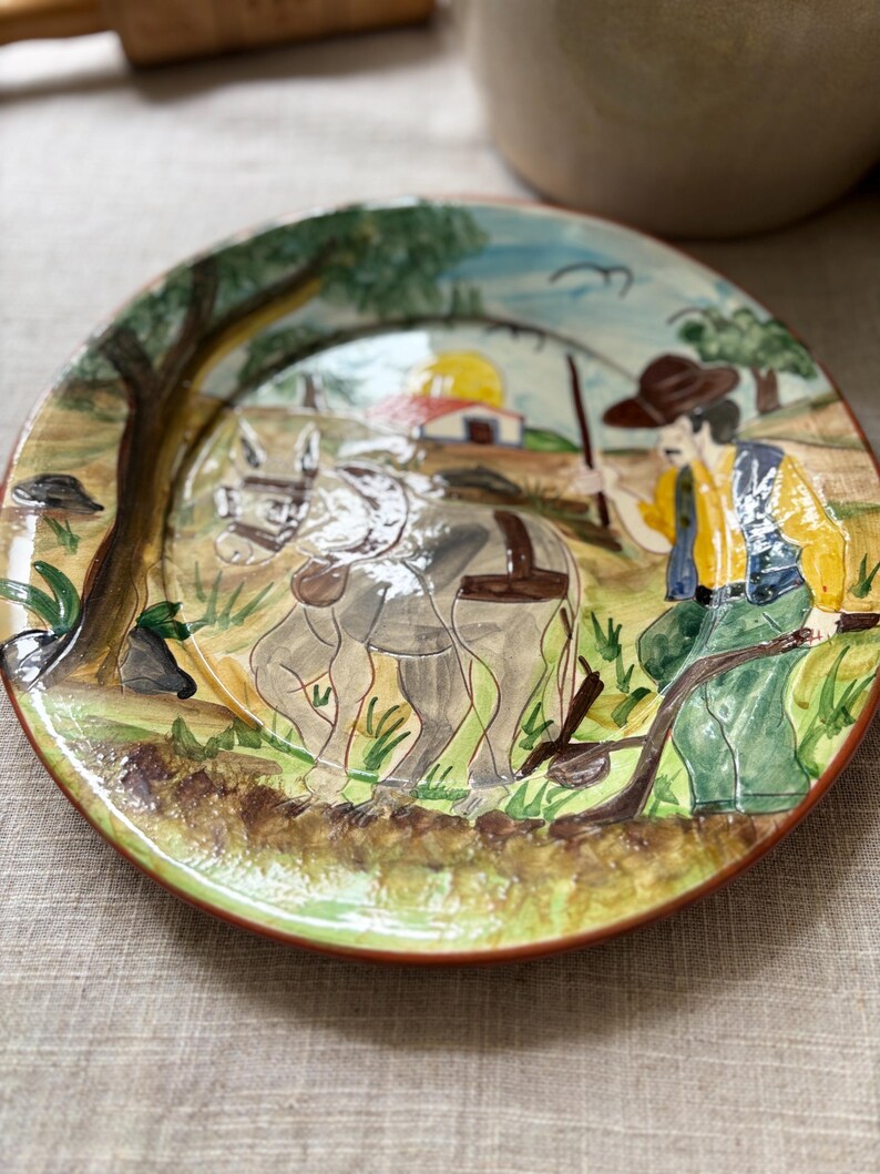 May include: A round ceramic plate featuring a hand-painted scene of a person in a brown hat and yellow shirt guiding a grey donkey through a field. The background includes a small building, a tree, and a yellow sun, all rendered in a glossy finish.