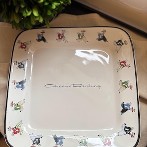 May include: A square white ceramic plate with a thin black rim. The border features a repeating pattern of illustrated figures in red, blue, and green clothing carrying trays. The center of the plate displays the black cursive text Cheers Darling.