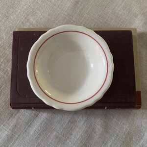 May include: A small, round, white ceramic bowl with a scalloped edge and a single thin red circular line accenting the interior. The bowl is positioned centrally on top of a rectangular, dark maroon-colored hardbound book with a visible spine.