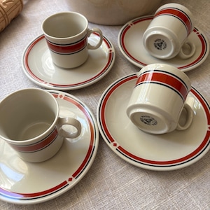 May include: Four off-white ceramic espresso cups and matching saucers featuring a horizontal red stripe bordered by thin black lines. The saucers include small red diamond accents. The undersides of the cups are marked with Made in China text.