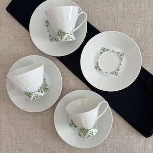 May include: Four white teacups and saucers with a green floral design. The cups have a flared shape and a handle. The saucers are round. The set is arranged on a neutral surface with a black cloth.