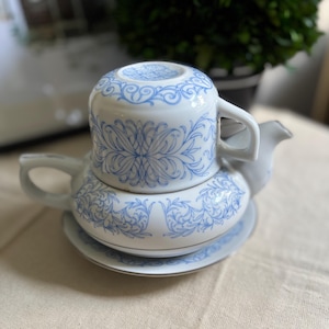 May include: A white ceramic tea set for one, consisting of a teapot, a teacup, and a saucer. The pieces feature a delicate light blue floral and scrollwork pattern. The teacup is stacked upside down on top of the teapot, which has a handle and spout.