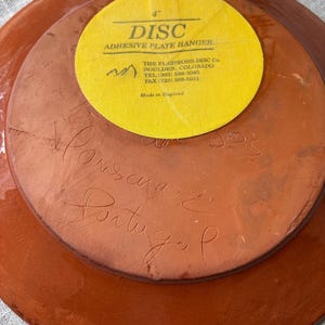 May include: The back of a round terracotta plate featuring a yellow adhesive plate hanger with the text 4" DISC ADHESIVE PLATE HANGER THE FLATIRONS DISC Co. BOULDER, COLORADO. Handwritten cursive text is etched into the clay surface reading Portugal.