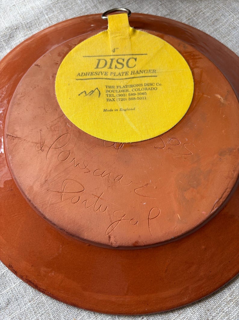 May include: The back of a round terracotta plate featuring a yellow adhesive plate hanger with the text 4" DISC ADHESIVE PLATE HANGER THE FLATIRONS DISC Co. BOULDER, COLORADO. Handwritten cursive text is etched into the clay surface reading Portugal.