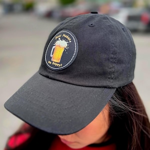 Może przedstawiać: Czarna czapka bejsbolowa z zakrzywionym daszkiem. Czapka posiada okrągłą naszywkę z grafiką kufla piwa i napisem "Beer Makes Me Happy!". Czapka wykonana jest z miękkiego materiału i nadaje się do noszenia na co dzień.