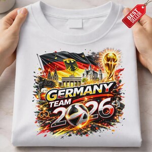 K&ouml;nnte beinhalten: Wei&szlig;es T-Shirt mit einem farbenfrohen Grafikdesign. Das Design beinhaltet die deutsche Flagge, einen Fu&szlig;ball, eine Troph&auml;e und den Text "GERMANY TEAM 2026". Das Shirt hat auch ein "BEST SELLER"-Etikett.