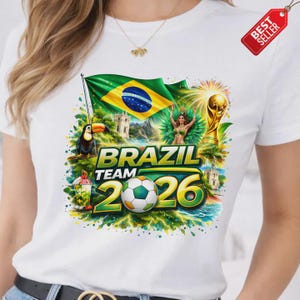 K&ouml;nnte beinhalten: Wei&szlig;es T-Shirt mit farbenfrohem Grafikdesign. Das Design zeigt die brasilianische Flagge, einen Tukan, einen Fu&szlig;ball und den Text "BRAZIL TEAM 2026". Ein "BEST SELLER"-Etikett ist angebracht.