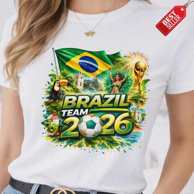 K&ouml;nnte beinhalten: Wei&szlig;es T-Shirt mit farbenfrohem Grafikdesign. Das Design zeigt die brasilianische Flagge, einen Tukan, einen Fu&szlig;ball und den Text "BRAZIL TEAM 2026". Ein "BEST SELLER"-Etikett ist angebracht.