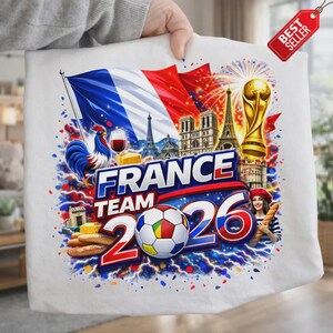 K&ouml;nnte beinhalten: Wei&szlig;es T-Shirt mit einem farbenfrohen Grafikdesign mit der Aufschrift "FRANCE TEAM 2026". Das Design zeigt die franz&ouml;sische Flagge, einen Fu&szlig;ball, den Eiffelturm, einen Hahn und eine Frau mit Baskenm&uuml;tze.