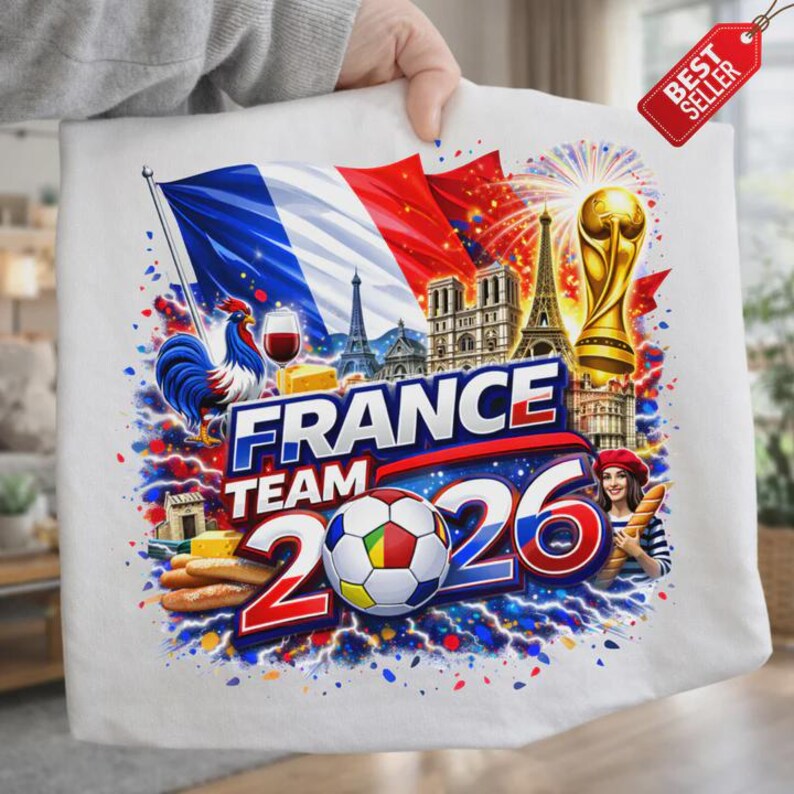 K&ouml;nnte beinhalten: Wei&szlig;es T-Shirt mit einem farbenfrohen Grafikdesign mit der Aufschrift "FRANCE TEAM 2026". Das Design zeigt die franz&ouml;sische Flagge, einen Fu&szlig;ball, den Eiffelturm, einen Hahn und eine Frau mit Baskenm&uuml;tze.