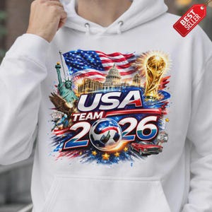 K&ouml;nnte beinhalten: Wei&szlig;er Kapuzenpullover mit einem grafischen Design mit dem Text "USA TEAM 2026". Das Design umfasst die amerikanische Flagge, die Freiheitsstatue, das Kapitol, einen Fu&szlig;ball und ein rotes Auto.