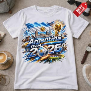 K&ouml;nnte beinhalten: Wei&szlig;es T-Shirt mit einem farbenfrohen Grafikdesign. Das Design zeigt den Text "Argentina TEAM 2026", einen Fu&szlig;ball, eine Flagge und eine Troph&auml;e. Das Design enth&auml;lt auch eine Stadtlandschaft und Figuren.