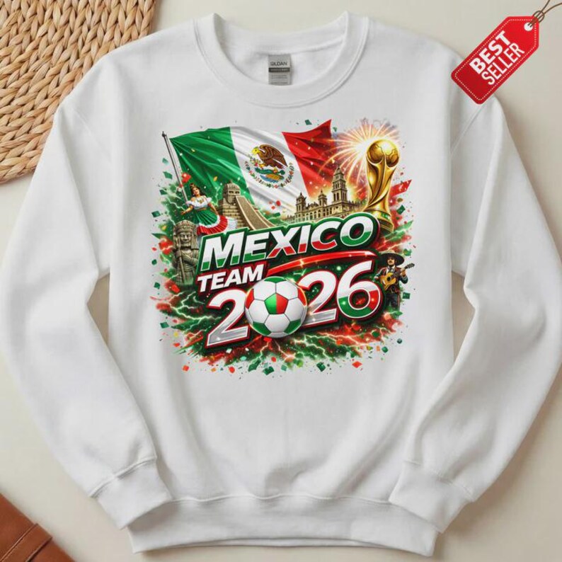 K&ouml;nnte beinhalten: Wei&szlig;es Sweatshirt mit einem farbenfrohen Grafikdesign f&uuml;r das Mexico Team 2026. Das Design beinhaltet die mexikanische Flagge, einen Fu&szlig;ball und den Text "MEXICO TEAM 2026". Ein "Bestseller"-Etikett ist angebracht.