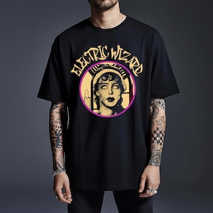 May include: A black short-sleeved t-shirt featuring a circular graphic on the chest. The design includes the text "ELECTRIC WIZARD" in yellow arched lettering above a stylised illustration of a person with pale eyes and a candle in a window.