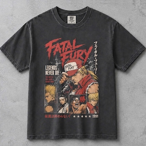 Puede incluir: Una camiseta gris carbón de manga corta con un estampado gráfico del videojuego Fatal Fury. El diseño incluye el texto Fatal Fury, Legends Never Die y caracteres japoneses, junto con ilustraciones de personajes y el año 1991.