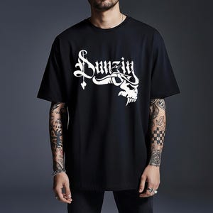 May include: A black short-sleeved t-shirt featuring a white graphic print on the chest. The design displays the word "Danzig" in a stylized gothic font, integrated with a skull illustration positioned beneath the lettering on the right side.