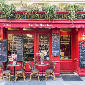 May include: A red shopfront with a sign that reads "Le Tir Bouchon" and an awning that reads "Le Tir Bouchon". The shopfront has two windows with black chalkboards displaying the menu. There are two tables with chairs outside the restaurant.
