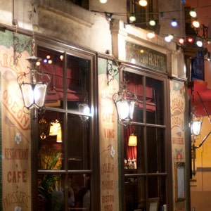 May include: A storefront with a sign that reads "Cafe Odeon Restaurant Cafe Patisserie" with string lights hanging above the entrance. The storefront has large windows with a red awning and a sign that reads "Vins".