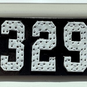 May include: A rectangular black sign featuring the white numbers 329. Each number is constructed from a white material with a series of small holes along the edges, through which white thread is stitched in a visible, repeating pattern.
