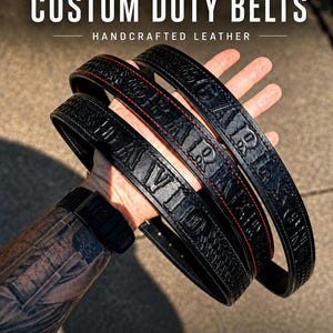 May include: Three black leather duty belts featuring embossed lettering and decorative patterns. The belts display the text CUSTOM DUTY BELTS HANDCRAFTED LEATHER at the top. Each belt has distinct stitching and personalised embossed names.