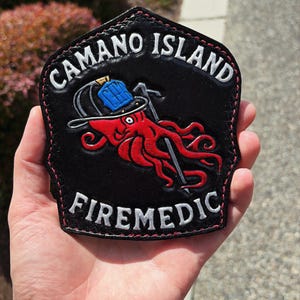 May include: A black leather patch with red stitching featuring a red octopus wearing a blue fire helmet and holding a silver tool. The text CAMANO ISLAND is arched at the top and FIREMEDIC is at the bottom, both in white embossed lettering.