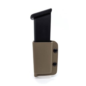 May include: A black firearm magazine inserted into a tan-colored molded holster. The holster features a textured surface and is secured with two black screws on the side. The magazine is rectangular with a slightly angled top edge.