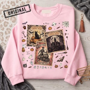 May include: A light pink sweatshirt featuring a graphic print of three vintage-style postage stamps labeled Victorian Halloween Curious Night, Ghost Train, and Night Ride Ticket, surrounded by small illustrations of ghosts, pumpkins, and bats.