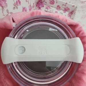 May include: A clear, round container with a white handle. The container is set on a pink surface, with a floral patterned background. The handle has a button and a logo. The container appears to be a food storage item.