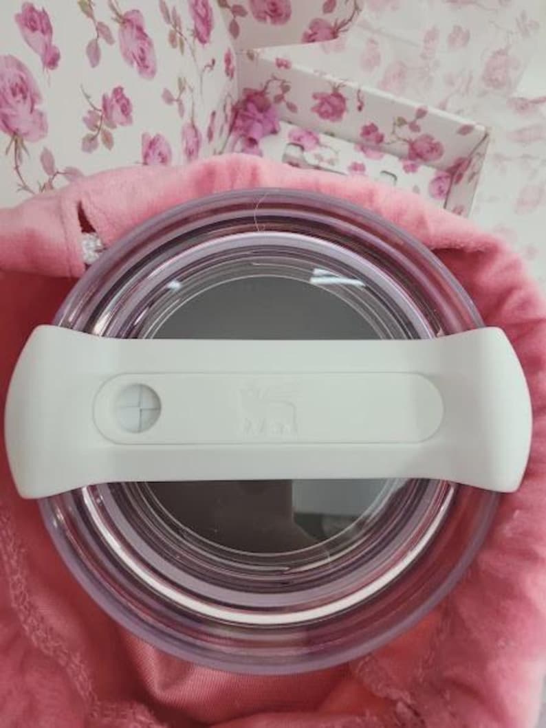 May include: A clear, round container with a white handle. The container is set on a pink surface, with a floral patterned background. The handle has a button and a logo. The container appears to be a food storage item.