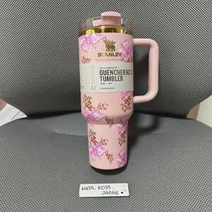 May include: A pink Stanley Quencher H2.0 tumbler with a floral design featuring pink bows and flowers. The tumbler has a handle and a clear lid. The text on the tumbler reads "THE FLOWSTATE QUENCHER H2.0 TUMBLER 40 OZ / 1.18L All Day Hydration".