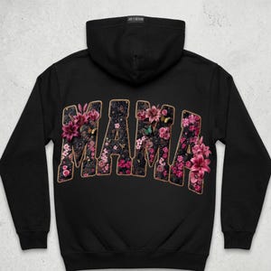 Pu&ograve; includere: Una felpa nera con cappuccio che presenta la parola MAMA in grandi lettere maiuscole arcuate sul retro. Le lettere sono riempite con un motivo scuro e strutturato di gigli rosa e viola, piccoli fiori e farfalle, con un bordo color sabbia.