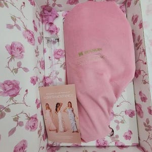May include: A pink Stanley Loveshackfancy insulated container inside a floral box. The box is decorated with pink roses on a white background. A small booklet with women in dresses is visible.