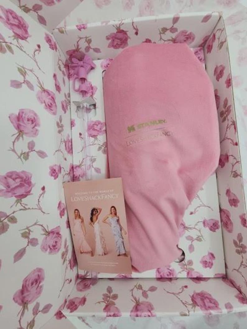 May include: A pink Stanley Loveshackfancy insulated container inside a floral box. The box is decorated with pink roses on a white background. A small booklet with women in dresses is visible.