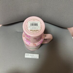May include: Pink Stanley travel mug with a floral pattern. The mug has a handle and a lid. The bottom of the mug has a sticker with the brand name and product information. The mug is decorated with pink bows and flowers.