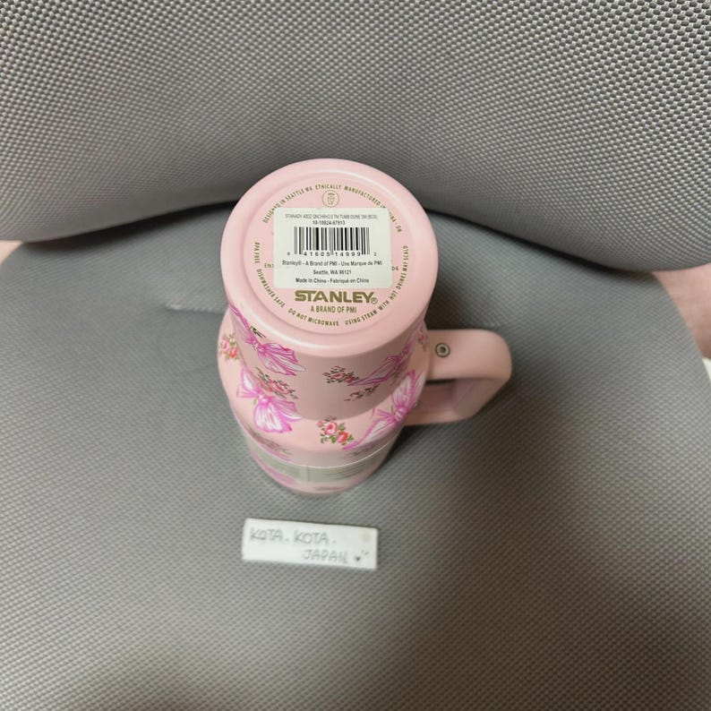 May include: Pink Stanley travel mug with a floral pattern. The mug has a handle and a lid. The bottom of the mug has a sticker with the brand name and product information. The mug is decorated with pink bows and flowers.