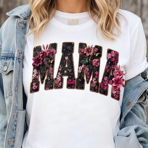 Pu&ograve; includere: Una t-shirt bianca a maniche corte con la parola MAMA in grandi lettere stampatello. Le lettere presentano uno sfondo scuro e testurizzato riempito con vari motivi floreali rosa e viola, inclusi gigli e piccoli fiori, con contorni dorati.