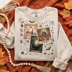 May include: A cream-coloured crewneck sweatshirt featuring a graphic print of three vintage-style postage stamps with Halloween themes, including a ghost writing at a desk, a pumpkin patch house, and a haunted forest, surrounded by small bats, ghosts, and webs.