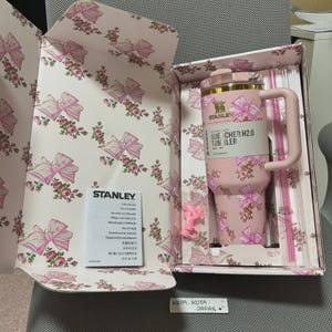 May include: A pink Stanley Quencher H2.0 tumbler with a floral and bow design, presented in a matching box. The tumbler has a handle and straw, and the box is decorated with bows and flowers. The box also contains a care and use booklet.