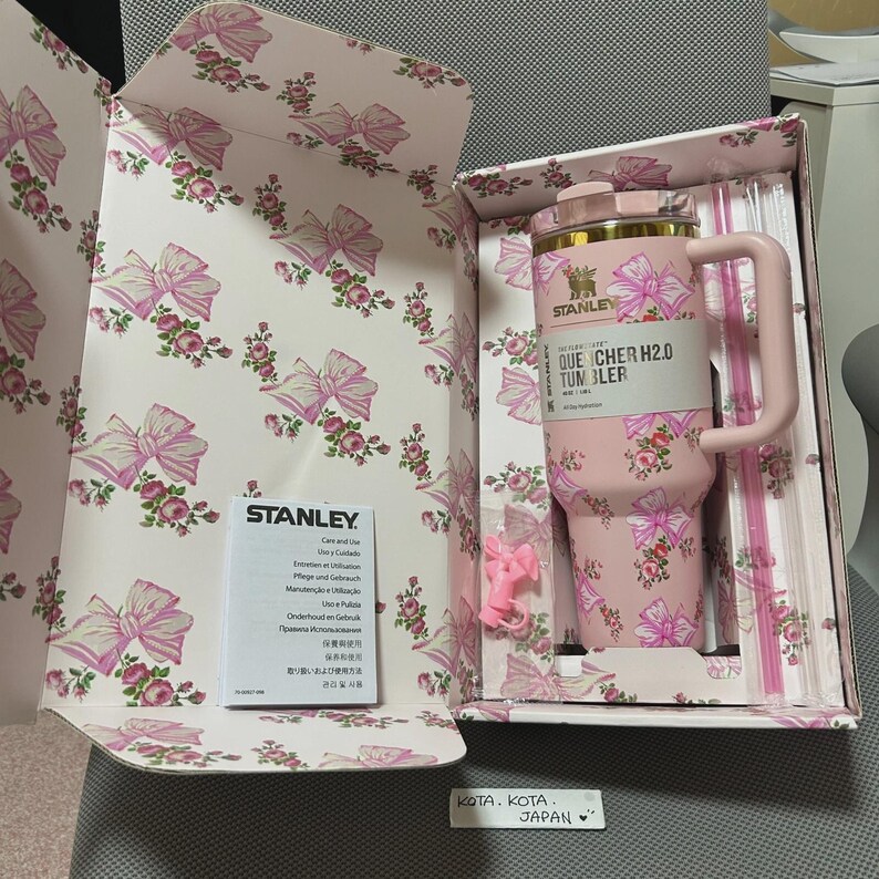May include: A pink Stanley Quencher H2.0 tumbler with a floral and bow design, presented in a matching box. The tumbler has a handle and straw, and the box is decorated with bows and flowers. The box also contains a care and use booklet.