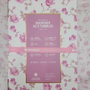 May include: A box with a floral rose pattern in pink and white. The box features a pink rectangle with the text "QUENCHER H2.0 TUMBLER" and the Stanley and Loveshackfancy logos. A pink ribbon wraps around the box.