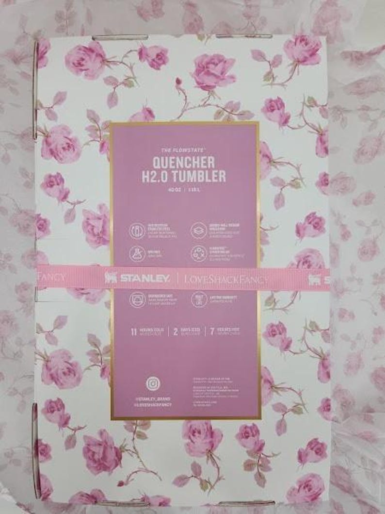 May include: A box with a floral rose pattern in pink and white. The box features a pink rectangle with the text "QUENCHER H2.0 TUMBLER" and the Stanley and Loveshackfancy logos. A pink ribbon wraps around the box.